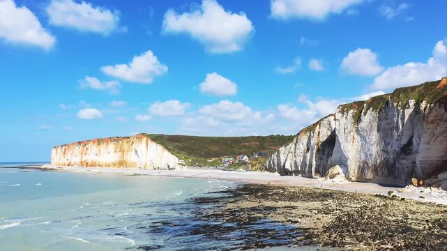 Scenic view of white chalk cliffs and rocky shoreline at Les Petites Dalles, Normandy, France under a bright blue sky with scattered clouds.