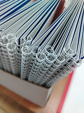 Stack of spiral-bound notebooks with white pages and blue covers arranged neatly in a box on a wooden surface, showcasing the binding and page edges