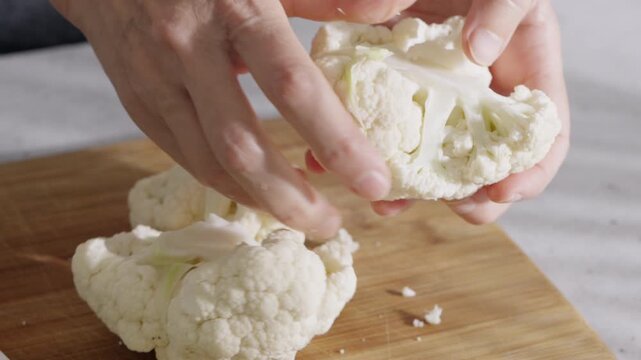 Close up of hands cutting cauliflower into small florets on a wooden board during kitchen food preparation.