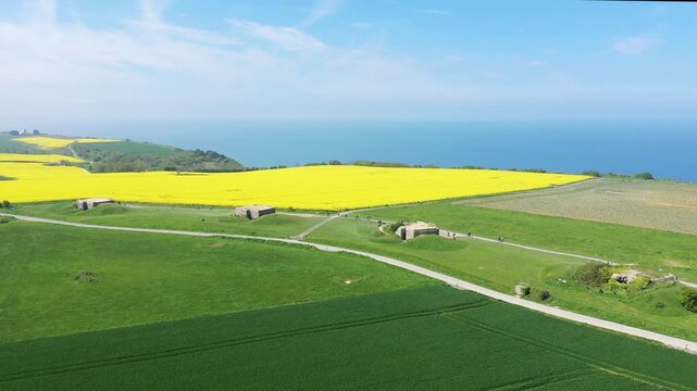 Aerial view of the Batterie de Longues sur Mer with green fields, yellow rapeseed, and the Normandy coast in the background under a clear blue sky.