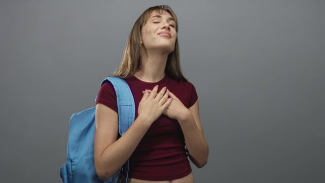 Young woman student with hands over chest and blue backpack in gray studio, eyes closed and grateful expression; gratitude reflection.