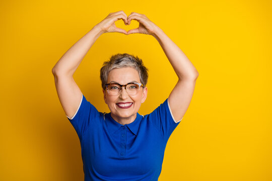 Smiling mature woman with short gray hair and glasses makes a heart shape with her arms in a blue polo against a bright yellow background conveying happiness and positive lifestyle energy