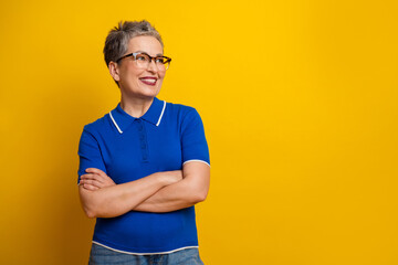 Senior woman in blue polo posing with arms crossed cheerful against yellow background