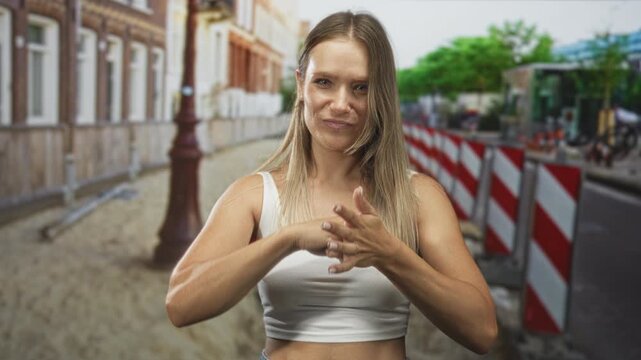 Woman clasping hands and grimacing on street beside construction barriers, rubbing knuckles and wearing white crop top; determination focus.