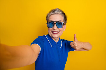 Aged mature female model in blue polo shirt wearing sunglass giving thumbs up against yellow...
