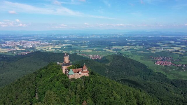 Scenic aerial shot of Chateau du Haut Koenigsbourg surrounded by lush green forest and expansive countryside in Alsace, France. Clear sky and panoramic landscape create a dramatic, historic atmosphere