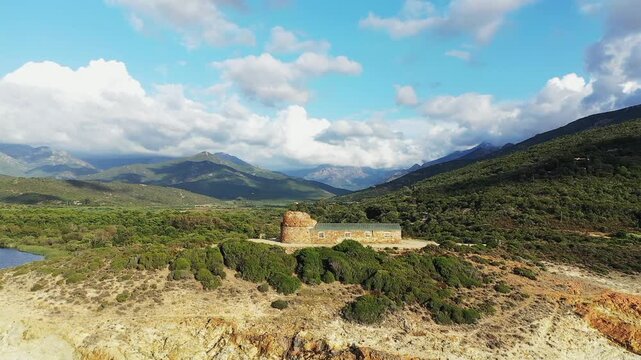 Aerial view of a historic stone tower surrounded by lush green hills and dramatic mountains in the Delta du Fango, Corsica. Scenic natural landscape with partly cloudy skies and rugged terrain.