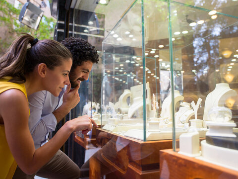 Couple window shopping for jewelry at a boutique display, smiling and pointing at rings and necklaces