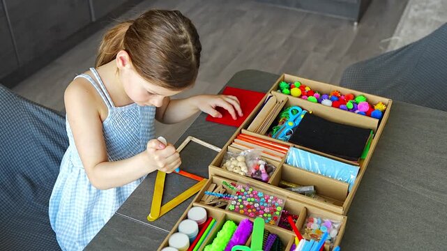 Little girl making craft from wooden sticks at table using colorful materials. Kids creativity fine motor skills development and hands on learning concept.