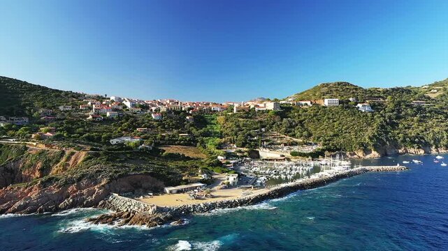 Scenic aerial shot of the marina and coastal village of Cargese, Corsica, surrounded by lush hills and the blue Mediterranean Sea under a clear sky.