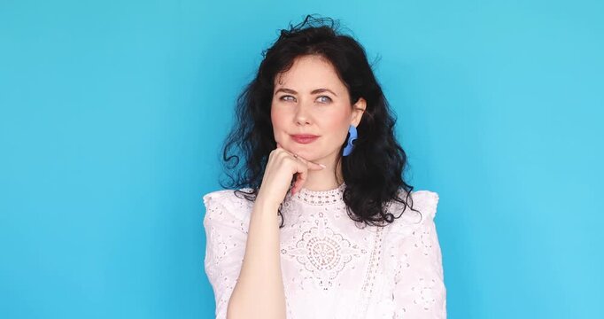 Thoughtful young woman in white lace dress thinking and finding an idea, holding chin then pointing finger up with eureka moment, looking excited and inspired on blue studio background