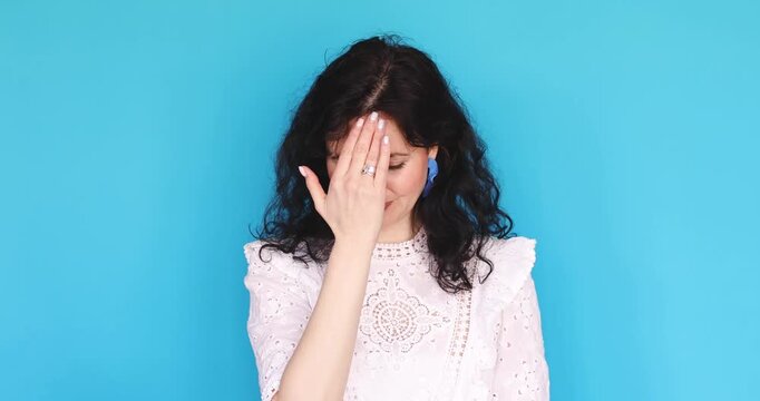 Young woman in white lace dress making facepalm gesture, covering eyes with hand and looking disappointed or embarrassed, feeling regret and shame isolated on blue studio background