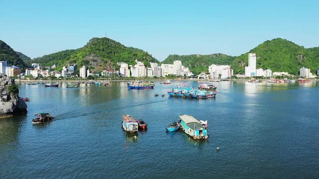 Colorful fishing boats float in the harbor against the backdrop of Cat Ba Island s cityscape and lush green hills under a clear blue sky.