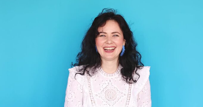 Young woman in white lace dress playfully and gently scolding someone with a smile, wagging her index finger in friendly admonishment against blue studio background