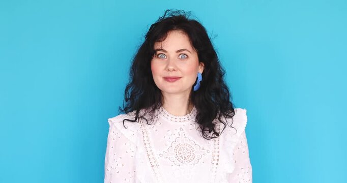Young woman with curly hair and blue earrings in white lace dress pointing two fingers at her blue eyes with "I'm watching you" gesture against blue studio background, looking at camera