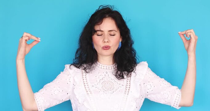 Peaceful young woman in white lace dress practicing meditation, breathing deeply with closed eyes and showing namaste mudra gesture then spreading hands in zen pose on blue studio background
