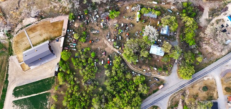 Aerial view of a junkyard dotted with colorful vehicles amidst lush greenery and buildings, contrasting with the adjacent open field, Palisade, Colorado, USA.