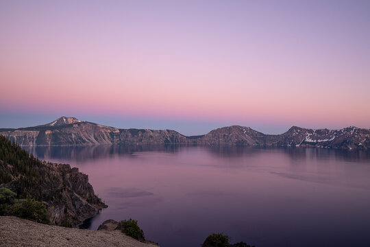 View of tranquil lake mirroring the soft, pastel hues of the twilight sky, framed by rugged cliffs and distant mountain peaks, Crater Lake, Oregon, United States.