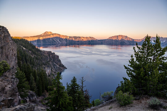 View of a serene blue lake reflecting the soft glow of the sunrise, surrounded by towering cliffs and lush green forests, Crater Lake, Oregon, United States.