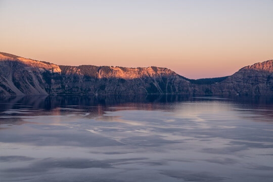 View of the serene lake reflecting the soft light of the sunset illuminating the rugged cliffs, creating a tranquil scene, Crater Lake, Oregon, United States.