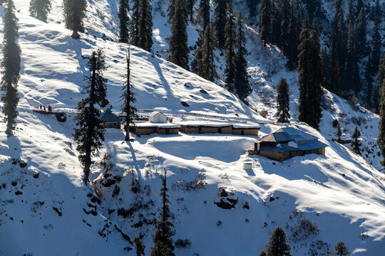View of snow-blanketed mountain slopes dotted with buildings and tall, dark trees under a bright sky, creating a serene winter landscape, Lasdana, Azad Kashmir, Pakistan.