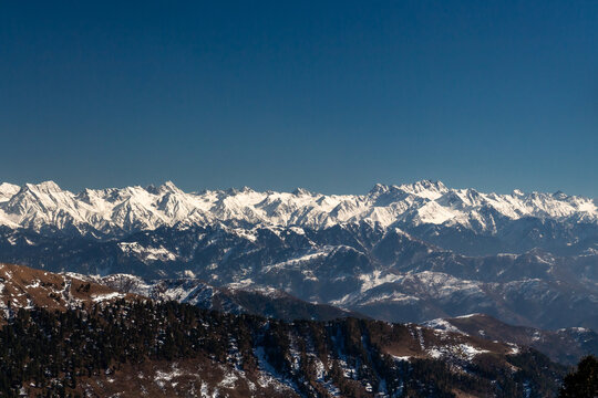 View of snow-capped mountains under a crisp blue sky create a stunning winter landscape, with dark green trees dotting the lower slopes, Lasdana, Azad Kashmir, Pakistan.