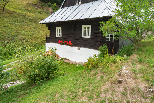 View of a charming dark wooden house with white trim and a silver roof sits nestled in a lush green hillside, Prasnica, Velka Fatra, Slovakia.