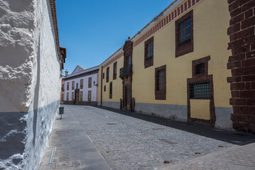Narrow cobblestone street in La Laguna, Tenerife, lined with historic buildings featuring colorful facades, wooden windows, and traditional Canarian architectural details under a clear blue sky © ihervas