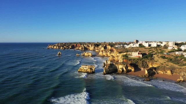Aerial view of rugged cliffs and sandy beaches along the Algarve coast near Lagos, Portugal, under clear blue skies. Picturesque seaside landscape with waves crashing against the rocks.