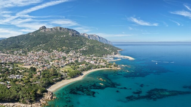 Aerial drone view of Santa Maria Navarrese with marina harbor, turquoise waters, sandy beach and dramatic limestone mountains along the eastern Sardinian coastline on a sunny day.