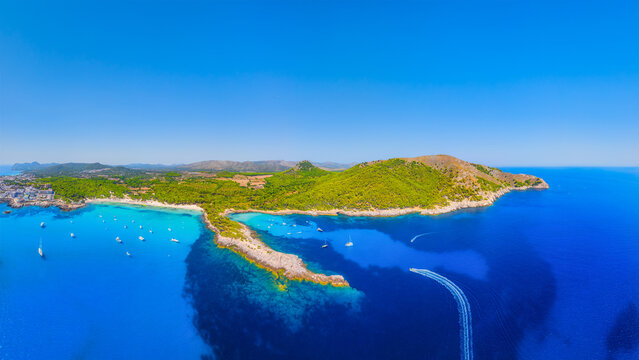 Aerial view of Cala Agulla beach and turquoise bay with boats in Mallorca. Scenic Balearic Islands coastline landscape in Spain. Summer vacation and Mediterranean nature from drone.