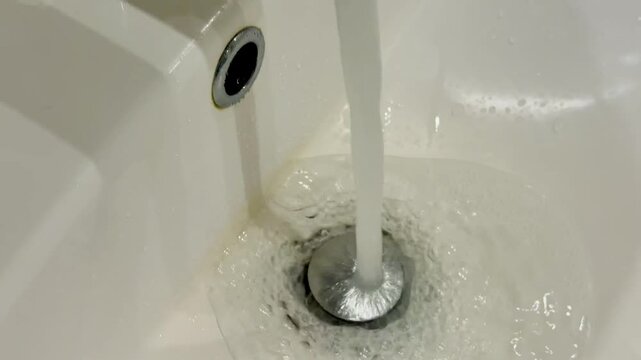 Dirty stainless faucet pouring water into unclean acrylic sink, visible mineral stains and soap scum around drain, slow steady.