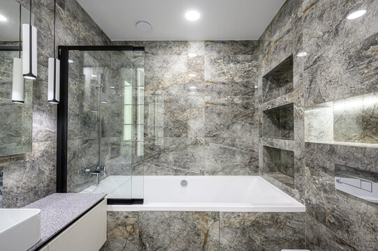 A modern bathroom with grey marble-patterned walls, white bathtub, glass shower screen with black frame, built-in niches, speckled countertop and sleek pendant lights, luxurious and minimalist.