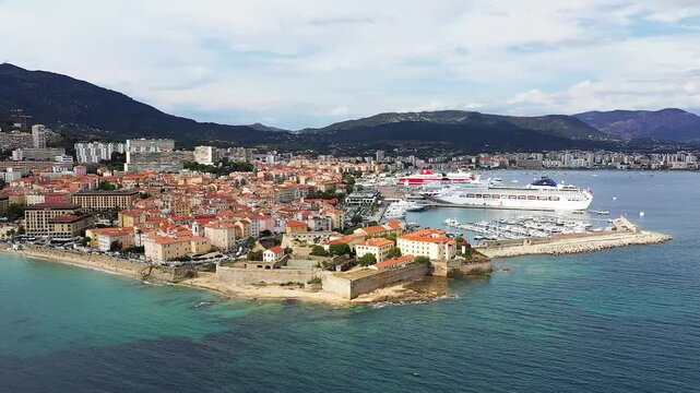 Scenic aerial view of Ajaccio with cruise ships docked at the harbor, historic buildings, and turquoise Mediterranean waters, set against a mountainous backdrop in Corsica, France.