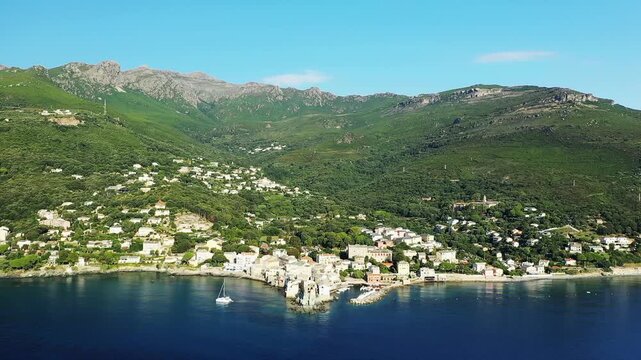 Scenic aerial view of Erbalunga village nestled on the Mediterranean coast with lush green hills and clear blue water, under a bright sky.