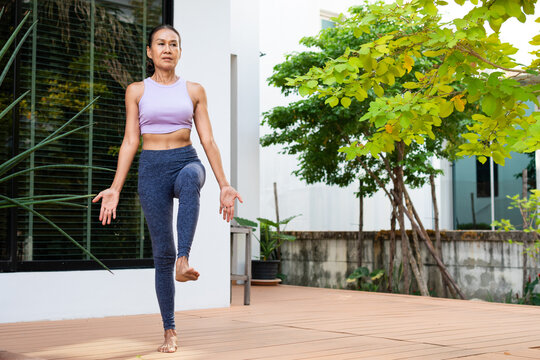 Senior woman age 62 practicing yoga outdoors standing on one leg with other bent, arms relaxed, symbolizing balance, wellness, healthy lifestyle, and authentic joyful everyday fitness and resilience