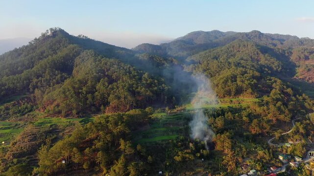 Aerial view of smoke rising from a forested mountain landscape near Sagada, Philippines, with terraced fields and rural scenery visible. Lush green hills and a clear sky create a dramatic natural