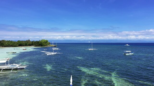 Traditional boats float on crystal clear blue water near a lush tropical shoreline in Panglao, Philippines under a bright sky.