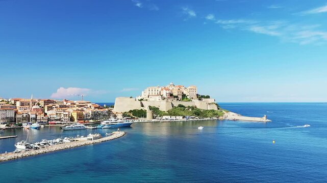 Scenic view of Calvi citadel and marina with yachts and boats on the clear blue Mediterranean Sea in Corsica, France under a bright sky.