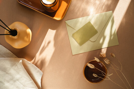 Overhead view of a blank business card on an envelope, a rustic vase with dried flowers, candle and napkin on a  table in sunlight