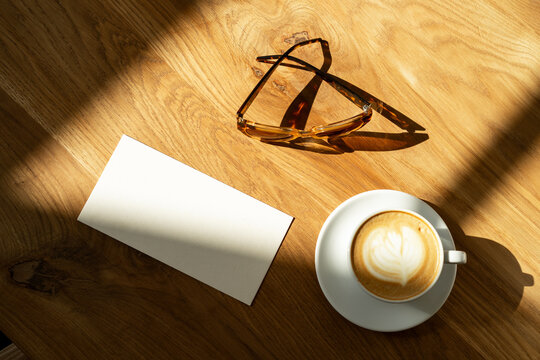 Overhead view of a cup of coffee, a pair of tortoiseshell sunglasses and a blank piece of card on a wooden table in sunlight