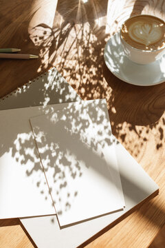 Close-up of a cup of coffee, blank pieces of paper and floral shadows  on a wooden table on a sunny morning