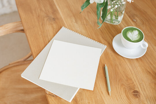 Close-up overhead view of a matcha latte hot drink, blank notepad and paper on a wooden table next to a bouquet of flowers in a vase