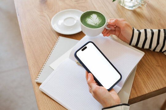 Close-up of a woman sitting in a cafe drinking a hot matcha latte while looking at a mobile phone with a blank screen