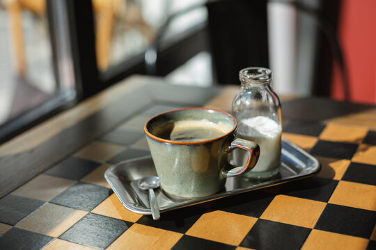 Close-up of a cup of black coffee and a small jug of milk on a metal tray on a checked table