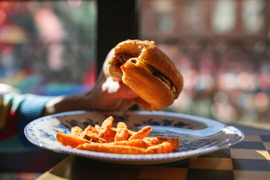 Close-up side view of a man sitting in a cafe on a sunny day eating a hamburger with sweet potato fries