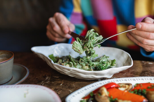 Close-up of a man sitting at a table eating a green salad with smoked stracciatella cheese and assorted roasted vegetables