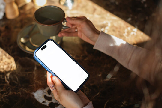 Close-up overhead view of a woman holding a mobile phone with a blank white screen and drinking a coffee