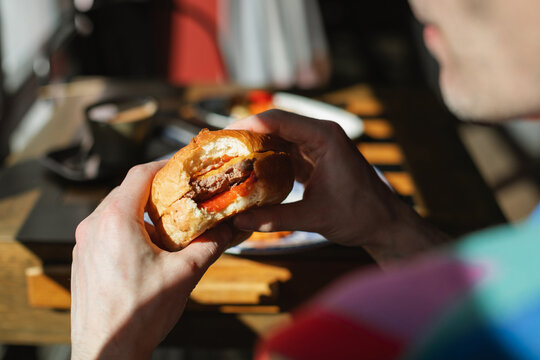 Close-up over the shoulder view of a man sitting at an outdoor cafe on a sunny day holding a half eaten hamburger