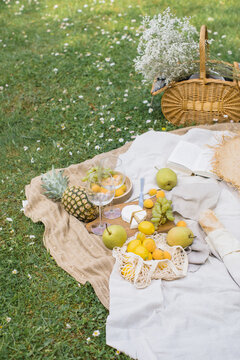 Close-up of a wicker basket with a bunch of baby's breath flowers, an open book, straw hat, fresh baguette, cheese, pineapple,  assorted fruit and wine glasses on a picnic blanket in a summer garden